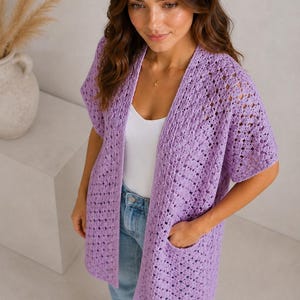 May include: A light purple, open-front crochet cardigan with short sleeves and two front pockets. The cardigan is made of a soft, open-weave material. The model is wearing a white tank top and blue jeans.