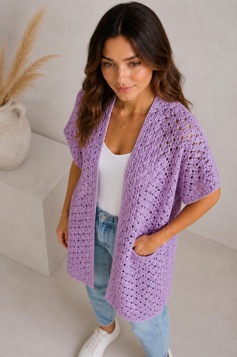 May include: A light purple, open-front crochet cardigan with short sleeves and two front pockets. The cardigan is made of a soft, open-weave material. The model is wearing a white tank top and blue jeans.