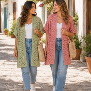 May include: Two women walking outdoors, each wearing a long, open-front cardigan. One cardigan is a soft green, the other is a dusty rose. Both are paired with light wash jeans, white tops, and straw tote bags. The cardigans have a delicate, open-knit design.