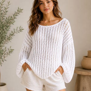 Pode incluir: Um suéter branco de malha aberta com mangas compridas e largas. O suéter tem um decote largo e é combinado com calções de cor clara. Os calções têm uma bainha desfiada. A imagem é tirada com luz natural.