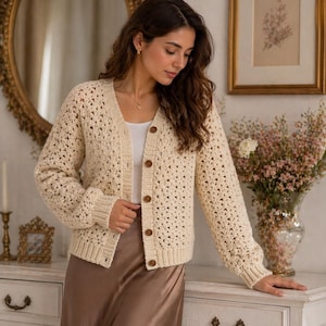 May include: Cream-colored, open-weave cardigan with long sleeves and wooden buttons. The cardigan is paired with a white top and a satin skirt. The image is set in a well-lit room with a mirror and floral arrangements.