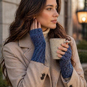 Puede incluir: Una persona con guantes sin dedos azules y un abrigo beige sostiene una taza de cerámica. Los guantes son de punto texturizado en tonos azules. El abrigo tiene un diseño clásico con botones oscuros. La taza está moteada con un asa pequeña.