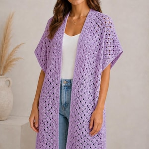May include: A long, open-front, lilac-colored crochet cardigan with short sleeves. The cardigan is worn over a white top and blue jeans. The crochet pattern is open, creating a textured look. The model is standing in front of a neutral background.