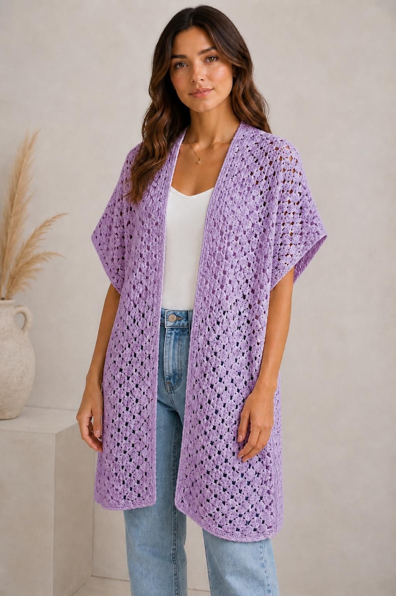 May include: A long, open-front, lilac-colored crochet cardigan with short sleeves. The cardigan is worn over a white top and blue jeans. The crochet pattern is open, creating a textured look. The model is standing in front of a neutral background.