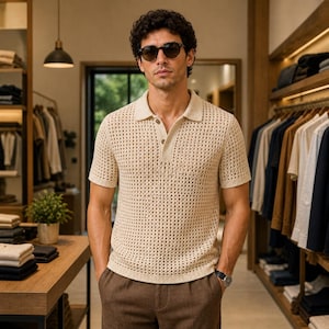 May include: A cream-coloured polo shirt with a textured, open-knit design. The shirt features a classic collar and buttoned placket. The model is wearing sunglasses and a watch. The background shows a clothing store with racks of apparel.