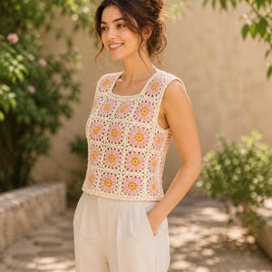 Op de afbeelding: Een crèmekleurige gehaakte top met vierkante halslijn en een bloemenpatroon in roze en geel. De mouwloze top wordt gecombineerd met een lichtgekleurde broek. De foto is buiten genomen in natuurlijk licht, met groen op de achtergrond.