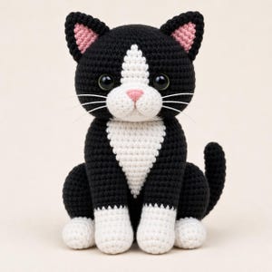 Könnte beinhalten: Handgehäkelte Katze in sitzender Position. Die Katze ist schwarz und weiß mit rosa Akzenten an Ohren und Nase. Sie hat große, schwarze Augen und weiße Schnurrhaare. Das ist ein weiches, plüschiges Spielzeug.