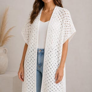 May include: A white, open-front crochet cardigan with short sleeves. The long, lightweight garment has a delicate, open weave design. The cardigan is worn over a white tank top and blue jeans. The overall style is casual and bohemian.