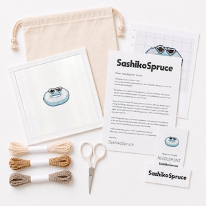 May include: A needlepoint kit featuring a square canvas with a blue donut graphic wearing sunglasses, three skeins of embroidery floss in beige, tan, and brown, white scissors, a beige drawstring bag, a pattern grid, and SashikoSpruce brand cards.