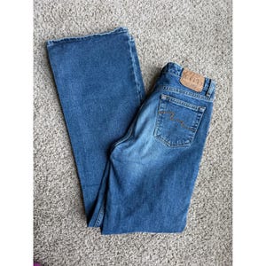 Puede incluir: Vaqueros azules de mezclilla con un diseño clásico de cinco bolsillos. Los vaqueros tienen una pernera acampanada y un lavado descolorido. Un parche de cuero marrón es visible en la cintura trasera.