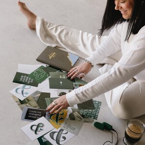 May include: A collection of cards with various designs and text, including the ampersand symbol and phrases like "I told my therapist about you." A journal and a cup of coffee are also present. The scene is set on a light-colored surface.
