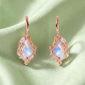 May include: Rose gold earrings with a leaf motif. Each earring has a rectangular, iridescent gemstone, complemented by small clear stones and intricate leaf-shaped details. The earrings are set against a soft green backdrop.