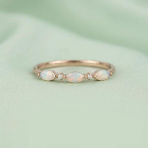 May include: A rose gold ring featuring three marquise-cut opals arranged horizontally, interspersed with two small round clear stones. The band is thin and polished, showcasing a delicate design that highlights the iridescent play of color in the opals.