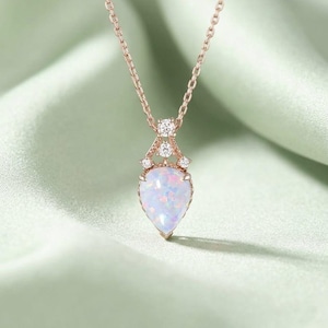 May include: A rose gold necklace featuring a pear-shaped opal pendant suspended from a delicate chain. The pendant is accented with three small, clear round-cut stones set in a decorative metal frame above the iridescent opal gemstone.