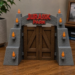May include: A grey, textured Jurassic Park gate replica featuring two brown wooden-style doors. The structure includes red lettering and eight small orange flame accents mounted on the sides. The design mimics a stone entrance with a central archway.