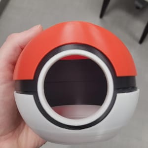 May include: A spherical container designed like a red, white, and black Pokeball. The object features a large circular opening on the front, revealing a hollow interior. The exterior surface is smooth with a distinct horizontal black band separating the top red hemisphere from the bottom white hemisphere.