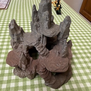 May include: A textured rock formation, possibly for aquarium decoration. The structure is a blend of grey and brown tones, with a cave-like opening and layered, organic shapes. The design mimics natural rock formations.