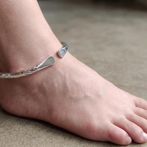 May include: A silver-colored, hammered-texture anklet. The open cuff design features a slightly flattened, rectangular shape at the ends. The anklet is worn on an ankle, showcasing its design and texture.
