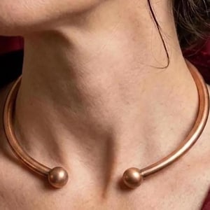 Puede incluir: Un collar de color cobre con un diseño liso y redondeado. El collar tiene un estilo sencillo y elegante con un extremo en forma de bola a cada lado. La joya se lleva alrededor del cuello.