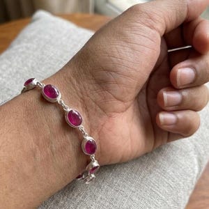 Può includere: Un bracciale in argento con pietre preziose ovali rosso intenso. Il bracciale è composto da maglie individuali, ciascuna con una pietra preziosa. Le pietre sono incastonate in argento. Il bracciale è mostrato su un polso.