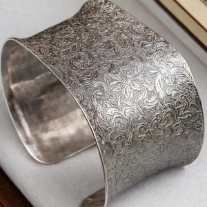 Peut inclure: Une tasse en céramique blanche avec une anse noire et le texte "But First, Coffee" dans un style manuscrit. La tasse est posée sur une surface blanche. La tasse mesure environ 10 cm de haut.