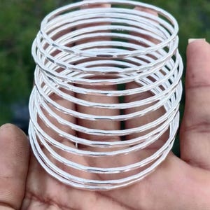 May include: A collection of silver bangles with a hammered finish. The bracelets are circular and have a shiny, reflective surface. The bangles are stacked, creating a layered effect. The background is blurred, with green hues.