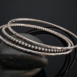 May include: Three silver bangles, each with a unique design, rest on a dark stone against a black background. One bangle is smooth, another has a hammered texture, and the third features small silver beads.