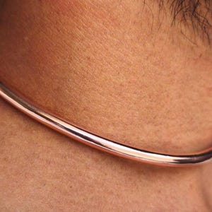May include: A polished copper-colored choker necklace. The necklace is a simple, smooth, and rounded band, worn around the neck. The image shows the necklace against a person's skin and a white shirt.