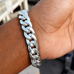 Puede incluir: Un brazalete de cadena de plata. El brazalete, hecho de eslabones entrelazados, se lleva en la muñeca. El brazalete brillante refleja la luz. Esta joya es un accesorio de moda.