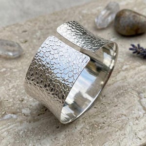 May include: A wide, open silver cuff bracelet with a textured, hammered surface. The bracelet has a rectangular shape with rounded edges and a gap for easy wear. The cuff is displayed on a light-colored stone surface.