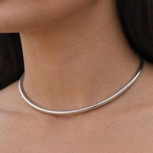 Puede incluir: Un collar gargantilla plateado y minimalista. El collar es una banda lisa y curvada que se ajusta cerca del cuello. La joya es simple y elegante, adecuada para el uso diario o ocasiones especiales.