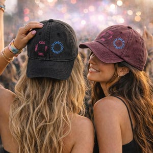 Könnte beinhalten: Zwei Baseballkappen, eine schwarze und eine weinrote, mit dem Text "Kiss the Time All Disco Occasionally" in einem kreisförmigen Design. Die Kappen werden von zwei Personen bei einer Outdoor-Veranstaltung getragen. Die schwarze Kappe hat rosa und blauen Text, und die weinrote Kappe hat rosa und blauen Text.