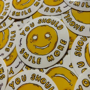 Può includere: Una collezione di adesivi rotondi con un design a faccina sorridente gialla. Gli adesivi hanno il testo "YOU SHOULD SMILE MORE" in un carattere giallo attorno alla faccina sorridente. Gli adesivi sono bianchi con un bordo giallo.