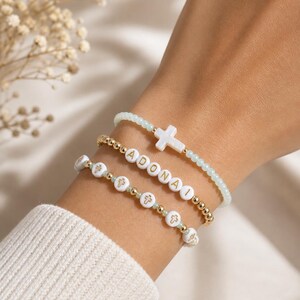 Peut inclure: Trois bracelets de perles. L'un est bleu clair avec une croix blanche. Un autre est doré avec le mot "ADONAI" épelé en lettres dorées. Le troisième bracelet est blanc avec des perles dorées et des croix blanches.