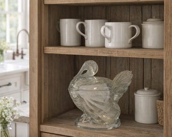 May include: A clear glass turkey-shaped lidded dish sits on a wooden shelf. The shelf is part of a wooden cabinet containing white ceramic mugs and canisters. The turkey dish features a textured surface and a detailed tail and neck design.