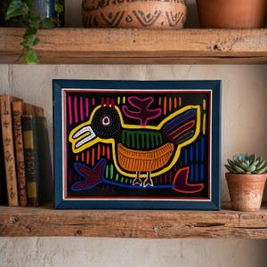 May include: Framed folk art depicting a colorful bird with a black background. The bird features yellow, orange, green, and blue details, framed in a navy blue wooden frame. The artwork is displayed on a rustic wooden shelf with books and a potted succulent.