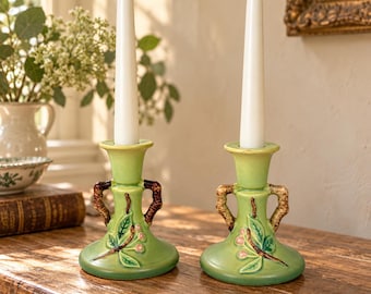 May include: A pair of light green ceramic candle holders with two handles each, featuring embossed leaf and berry motifs in brown and pink. Each holder supports a tall, slender, white taper candle, creating a balanced display.