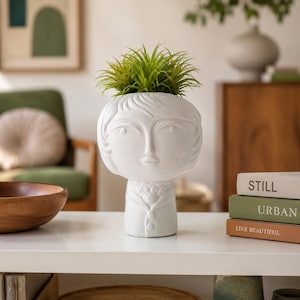 May include: A white ceramic planter shaped like a human head with a stylized face and a collared shirt design. The planter holds a vibrant green spiky plant. The surface of the planter is matte white, featuring sculpted facial features.