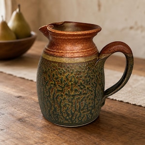 May include: A ceramic pitcher featuring a rounded body with a textured, mottled green and brown glaze. The upper section and the curved handle are finished in a smooth, solid reddish-brown glaze. The pitcher has a wide, flared rim and a spout.