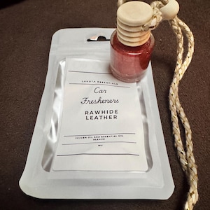 May include: A small glass bottle containing red liquid with a wooden cap and a braided cord, resting on a white pouch labeled Lakota Essentials Car Fresheners Rawhide Leather Jojoba Oil and Essential Oil Blends 8ml.