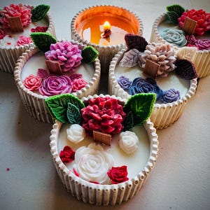 May include: A collection of decorative candles with floral designs. The candles are in white, ribbed containers and topped with colorful, sculpted flowers and green leaves. One candle is lit, showing a warm glow.
