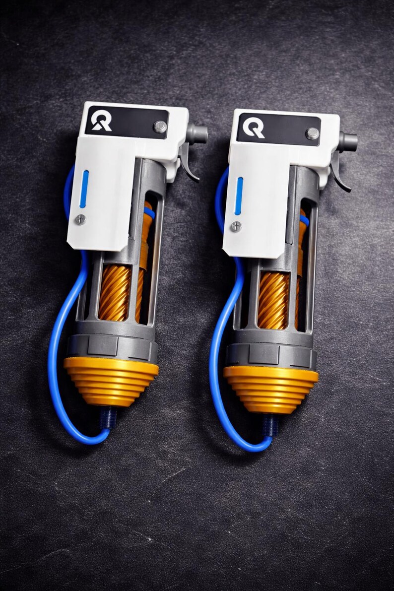 May include: Two handheld devices featuring a white top section with a black square containing a white letter Q, a grey frame, a central gold-colored spiral component, a yellow ribbed base, and a blue cable connecting the top to the bottom.