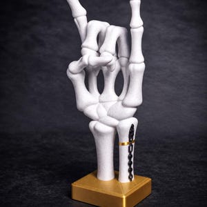 May include: A white speckled skeleton hand sculpture posed in a rock and roll gesture. The wrist features a black metal plate with screws and a thin gold band. The hand is mounted on a square, metallic gold base with a textured top surface.