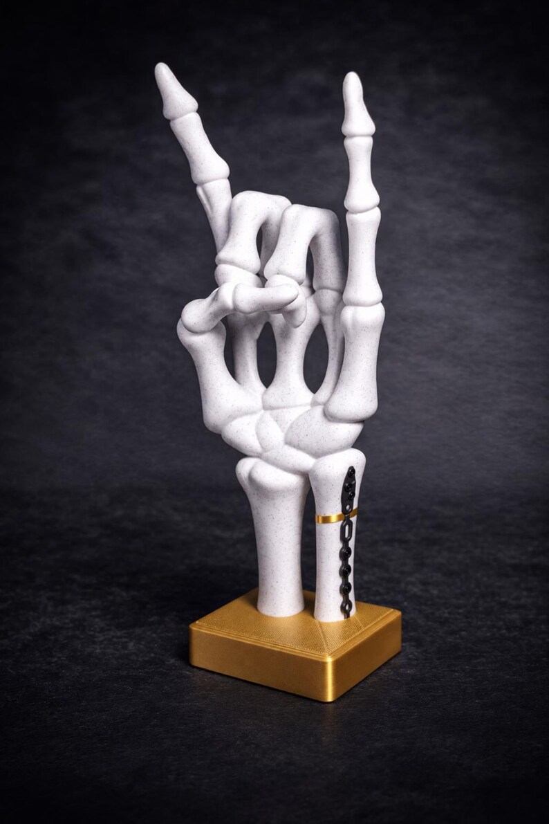 May include: A white speckled skeleton hand sculpture posed in a rock and roll gesture. The wrist features a black metal plate with screws and a thin gold band. The hand is mounted on a square, metallic gold base with a textured top surface.