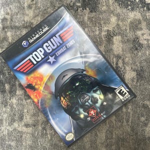 May include: A Nintendo GameCube video game case for Top Gun Combat Zones. The cover features a pilot wearing a helmet with a digital heads-up display, an explosion in the background, the game title in white and red text, and an ESRB Everyone rating.
