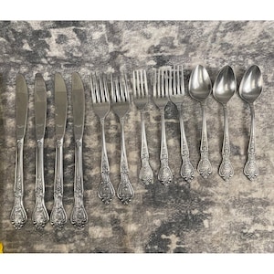 May include: A collection of vintage silverware, including knives, forks, and spoons, arranged on a patterned surface. The silverware has ornate floral detailing on the handles. The knives, forks, and spoons are made of a shiny, silver-colored metal.