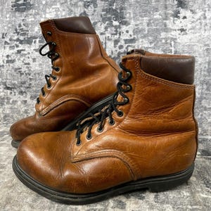 May include: A pair of brown leather work boots with black laces and a black sole. The boots have a worn appearance, suggesting they are well-used. The boots have a padded collar and a classic work boot design.