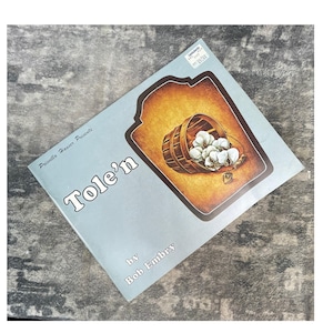 May include: A softcover book titled Tole'n by Bob Embry, presented by Priscilla Hauser. The light blue cover features an illustration of a wooden bucket tipped on its side, spilling out white cotton bolls against a dark orange background.