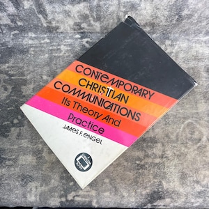 May include: A hardcover book titled Contemporary Christian Communications Its Theory And Practice by James F. Engel. The cover features a black top section, an orange middle band, a pink lower band, and a white base with a circular logo.