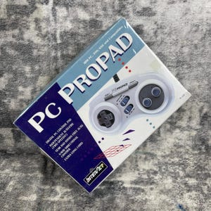 May include: A boxed PC Propad controller for IBM PC 386/486 and compatibles. The box is white and blue with the words "PC PROPAD" in large letters. The controller is white with gray buttons and a directional pad. The box also says "Interact".